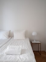 A bedroom with two beds, white bedding, a white lamp on a wooden nightstand, and a wooden floor.