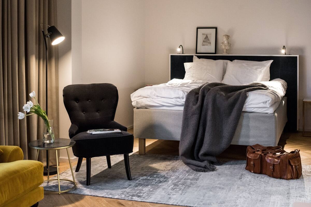 Ein Hotelzimmer mit Bett, Stuhl, kleinem Tisch, Lampe und Blumen in einer Vase.