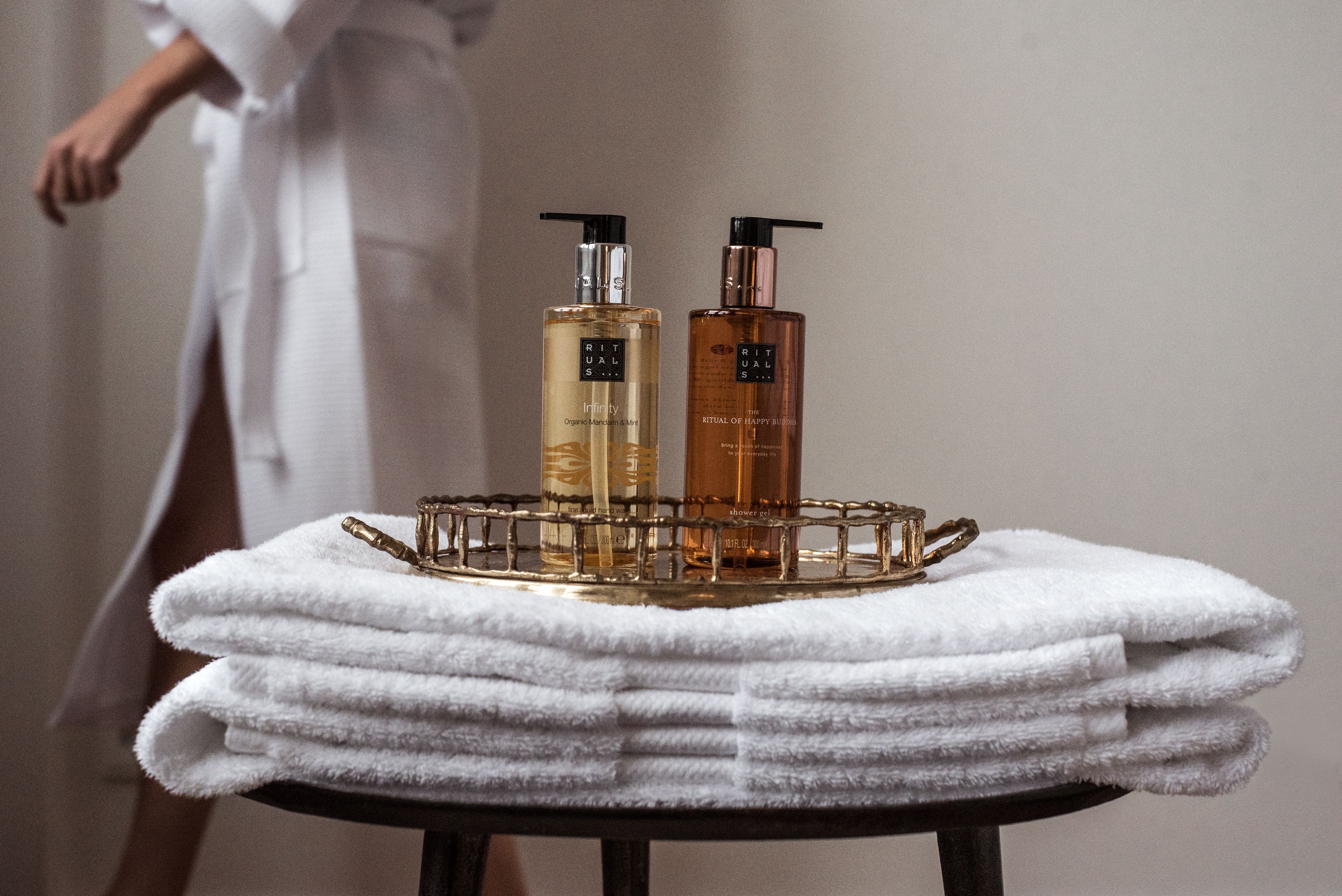 A bathroom with two bottles of Ritual Spa products on a tray, a stack of white towels, and a person in a robe in the background.