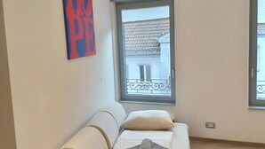 Un salon moderne avec un canapé beige, une fenêtre donnant sur un immeuble et une affiche « ESPOIR » accrochée au mur.
