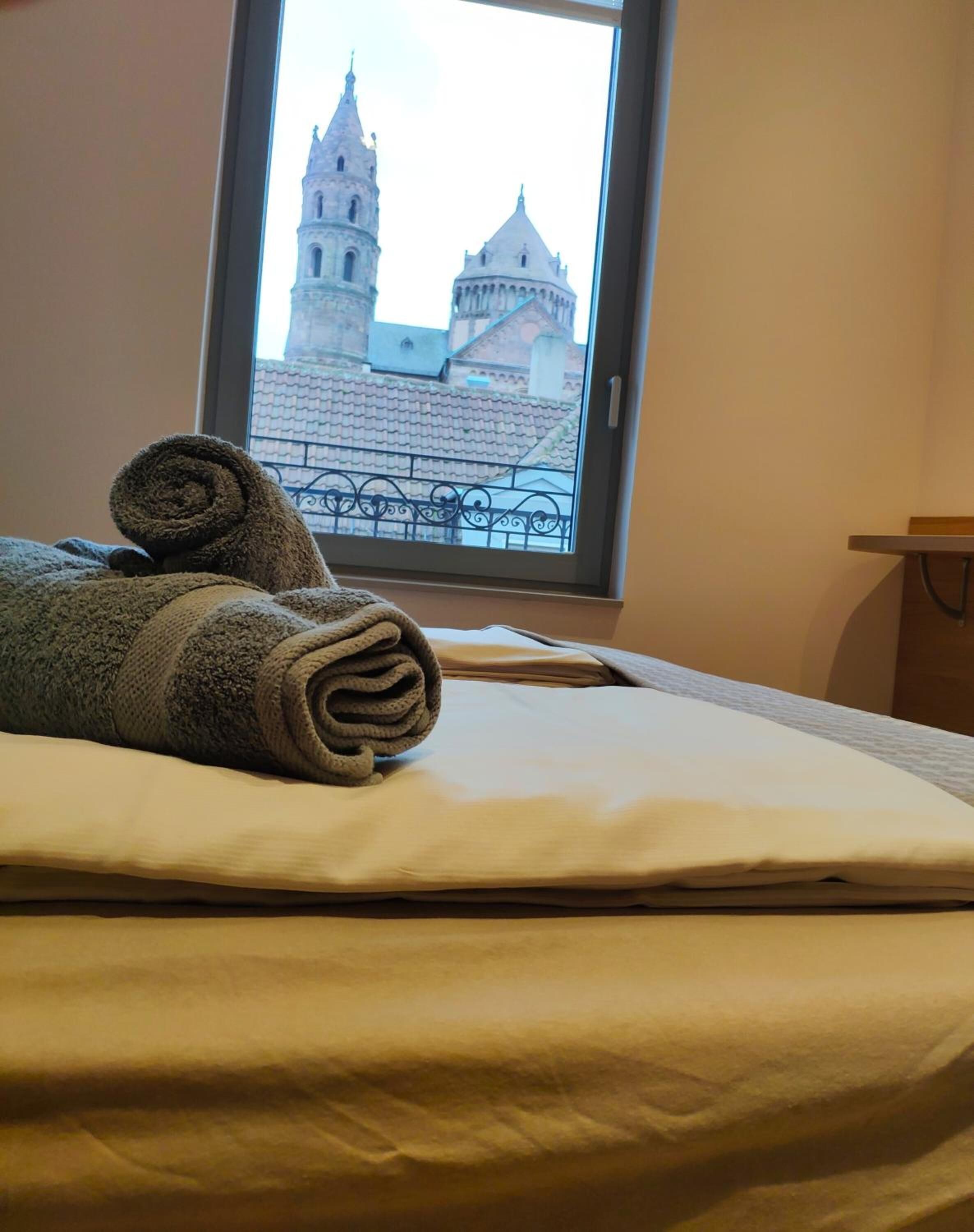 A neatly made bed with a rolled towel and a view of a historic tower through a window.