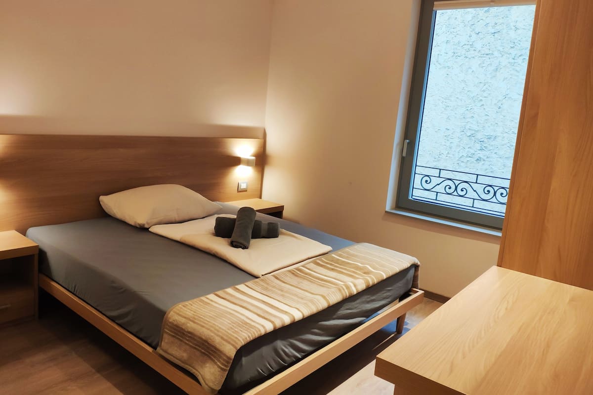 Una cama individual con cabecera de madera, una mesita de noche y otra mesita auxiliar. Se ve una ventana con vistas a un paisaje nevado.