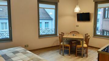 A room with a wooden dining table and chairs, a television mounted on the wall, and large windows with balconies.