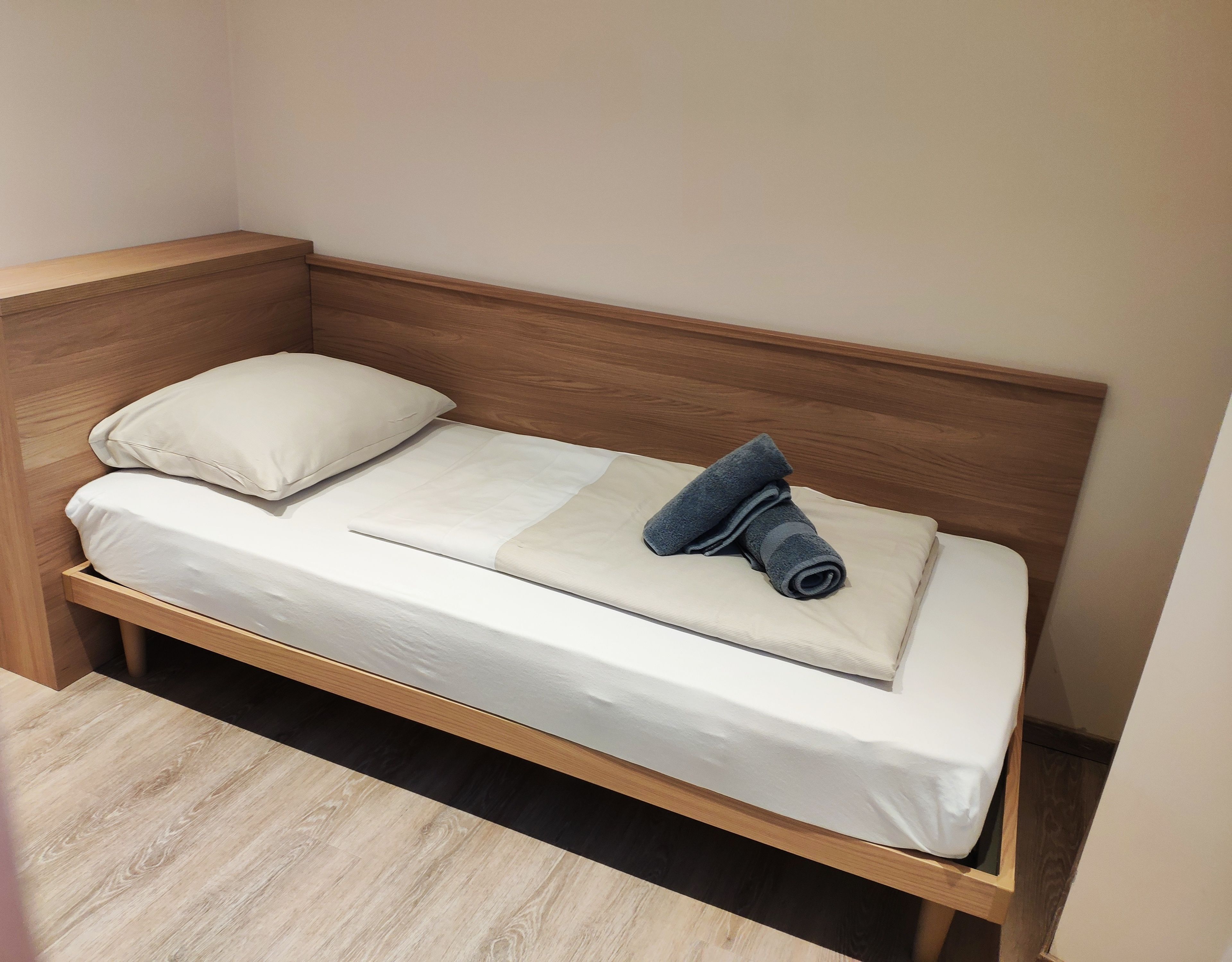 Una cama individual con estructura de madera, ropa de cama blanca y una toalla enrollada encima.