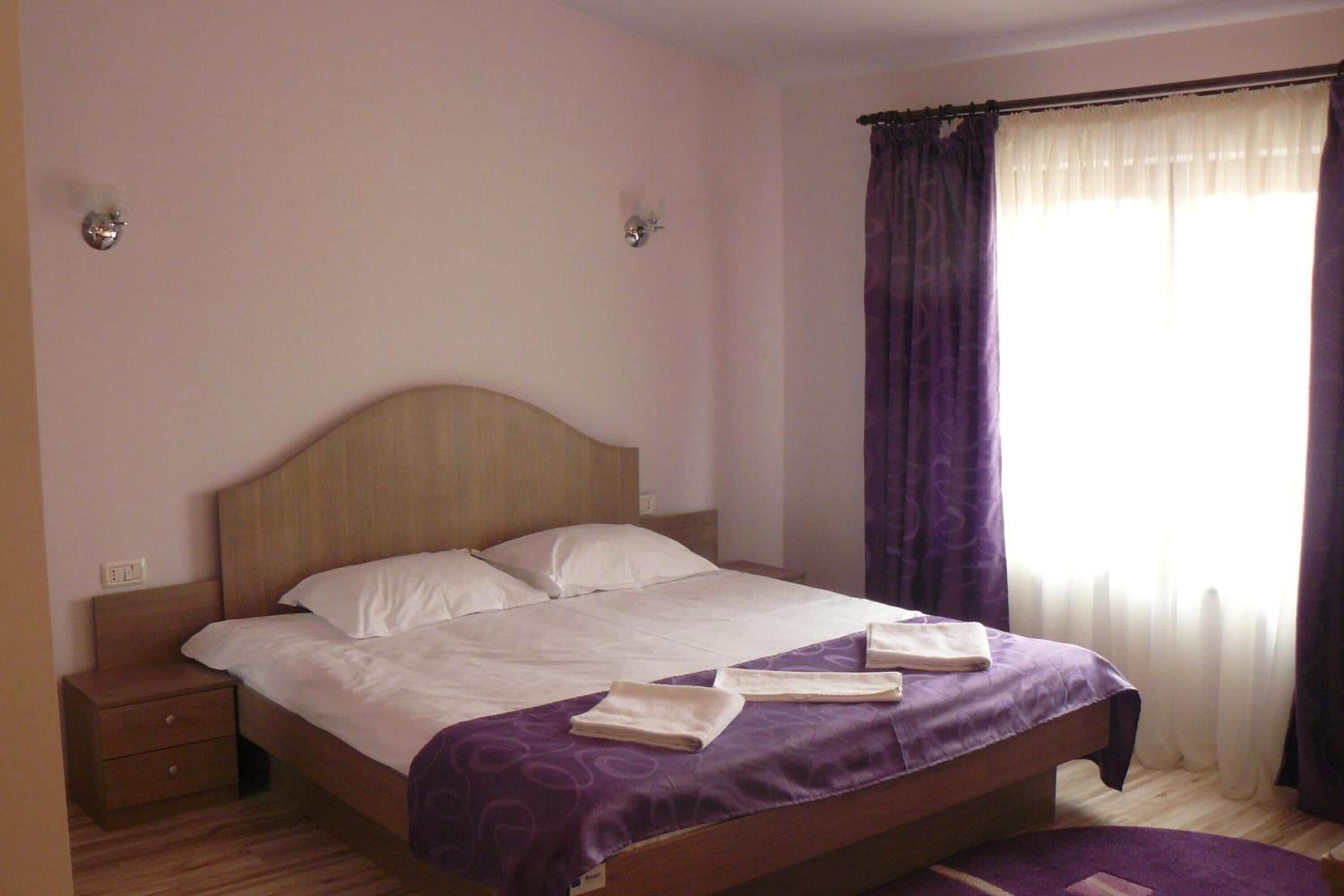 A neatly made bed with a wooden headboard, a bedside table with a drawer, and a window with purple curtains.