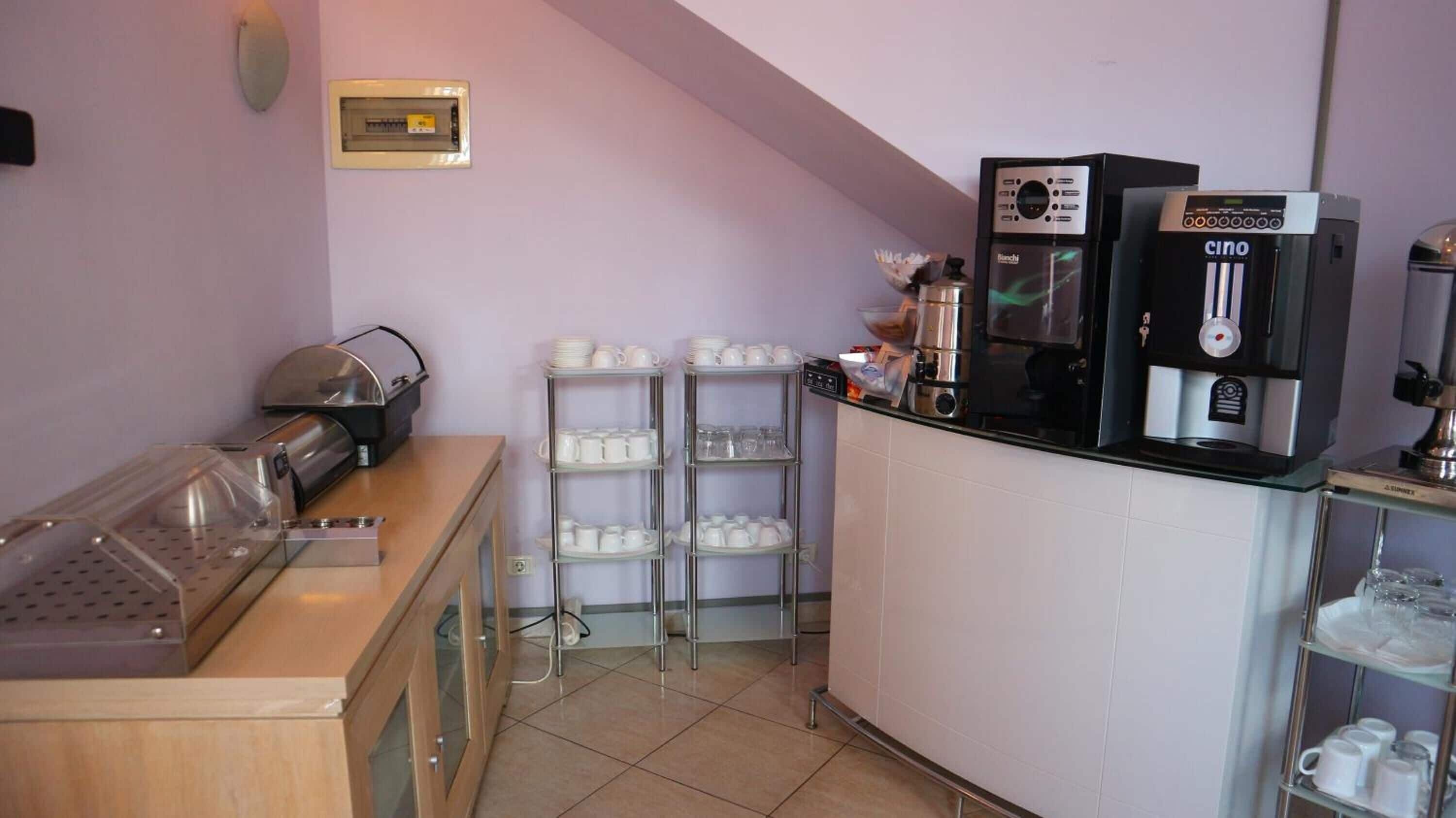 Una kitchenette equipada con cafetera, tostadora y dispensador de agua. Hay repisas con tazas y un refrigerador pequeño.