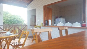 A wooden dining table with chairs, a kitchen area with a refrigerator and a coffee machine, and a view of trees outside. - Pousada Delicia de Sono (Cajueiro da Praia)