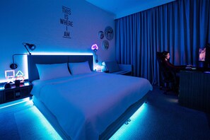 A hotel room with a bed, a desk, and a computer setup. There is a wall with a sign that says "THIS IS WHERE I AM."