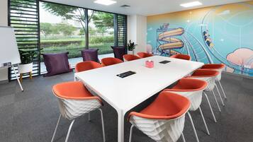 Uma sala de conferências com uma mesa branca, cadeiras laranja e brancas, um quadro branco e um mural de uma montanha-russa.