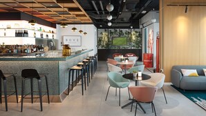 A modern bar area with a counter, bar stools, and a variety of bottles. There are several tables with chairs, a sofa, and a TV screen displaying a sports game. - Rove At The Park (Dubai)