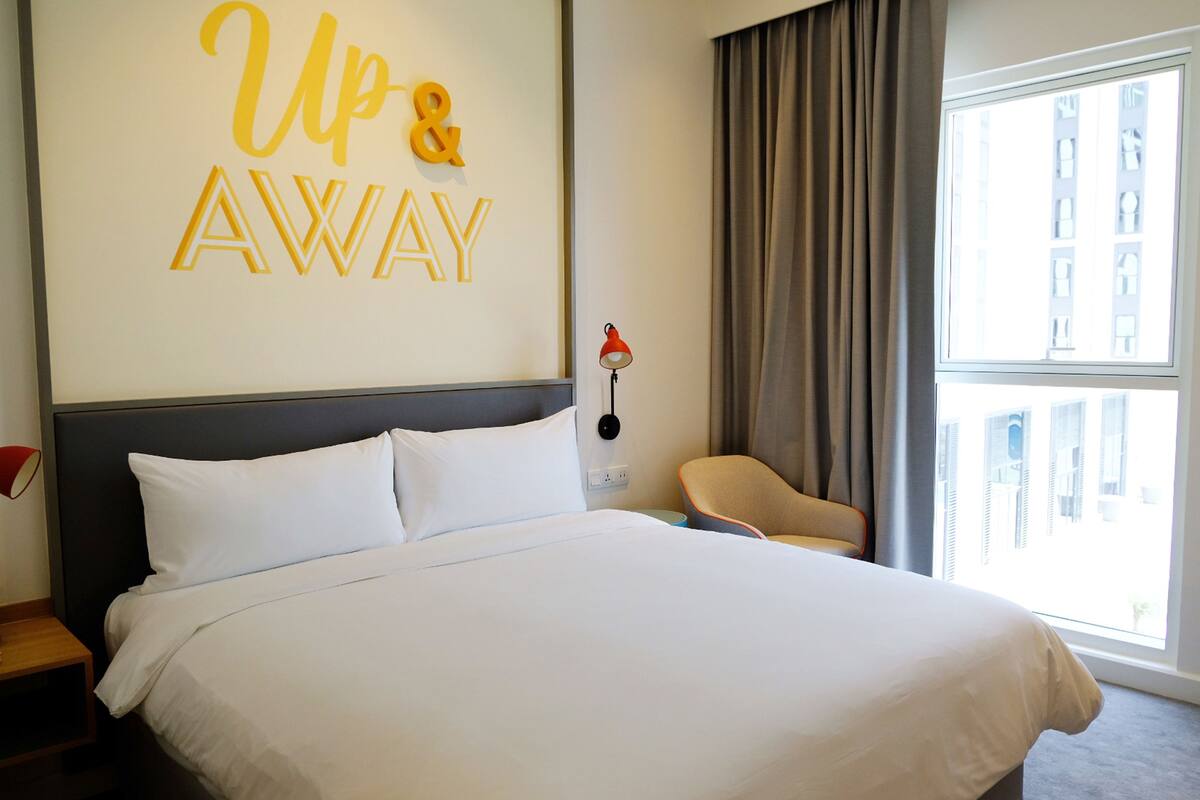 A hotel room with a bed, a chair, a lamp, and a large window.