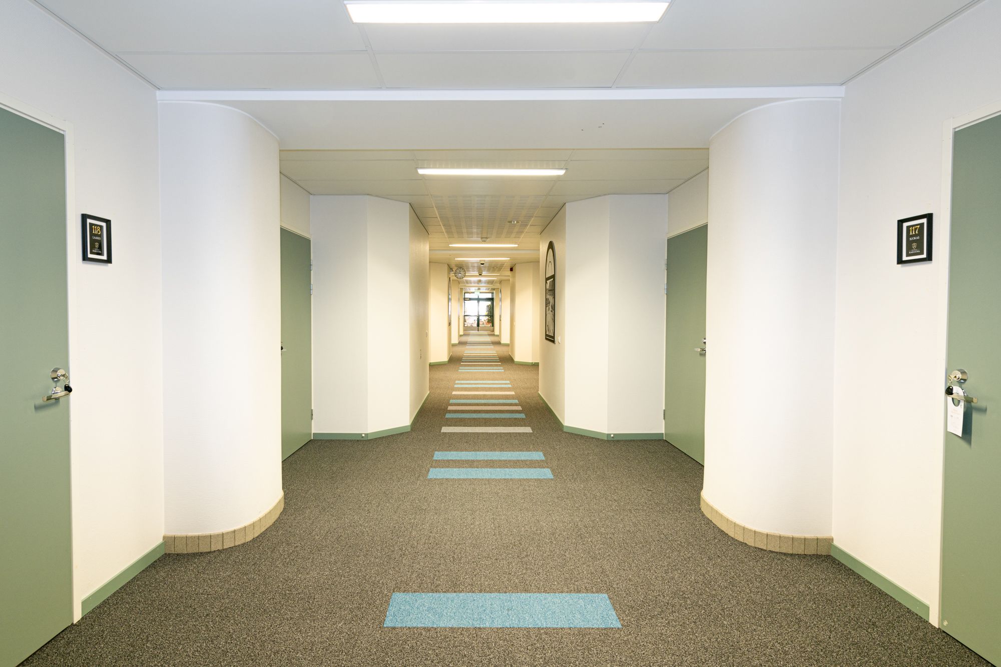 Ein Hotelgang mit Teppichboden, weißen Wänden und Türen auf beiden Seiten. Blaue Linien auf dem Boden zeigen einen Weg.