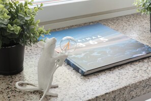 A white mouse figurine holding a magnifying glass on a kitchen counter with a book titled "AIMAA 365" and a potted plant.