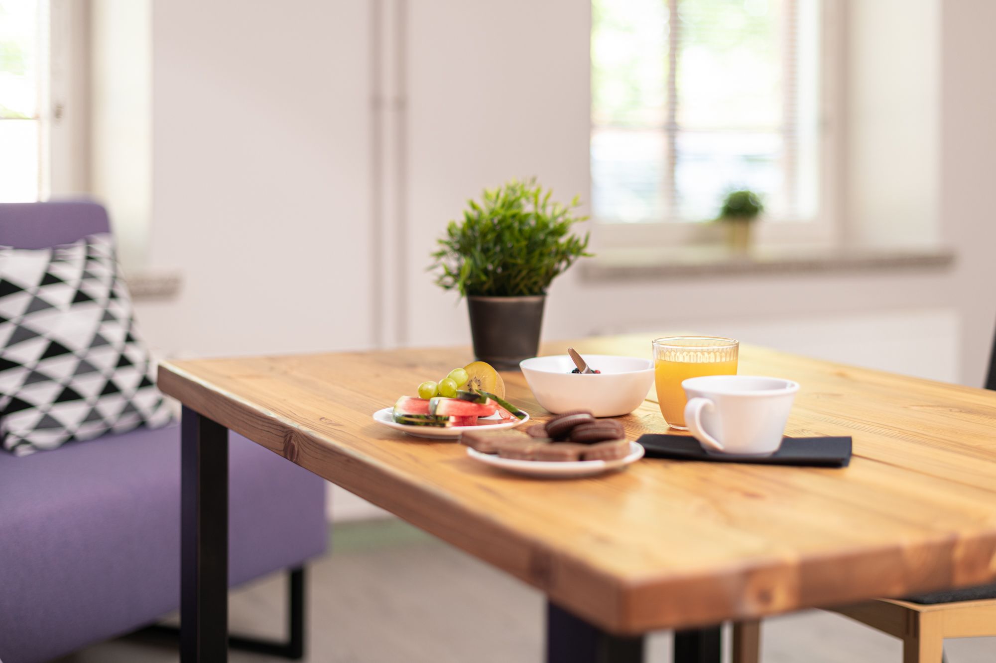 Une table en bois avec un bol de céréales, un verre de jus d’orange et une tasse. Une plante en pot se trouve à l’arrière-plan.