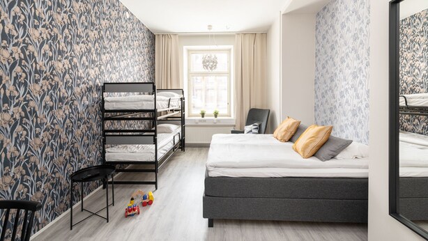 Ein Schlafzimmer mit Etagenbett, Schlafsessel, Schreibtisch und Stuhl. Ein Fenster mit Vorhängen und eine Pflanze im Fensterbrett.