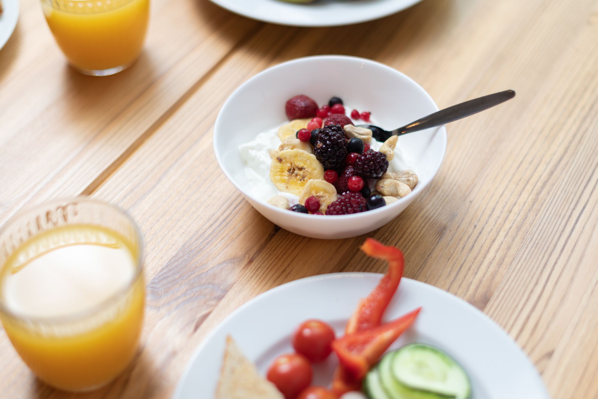 A breakfast setting with a bowl of yogurt topped with fruits and nuts, a glass of orange juice, and a plate with sliced vegetables.