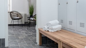 Ein sauberer, moderner Umkleideraum mit grauen Schließfächern, einer Holzbank mit aufgerollten Handtüchern und zwei Korbstühlen neben einem Heizkörper.