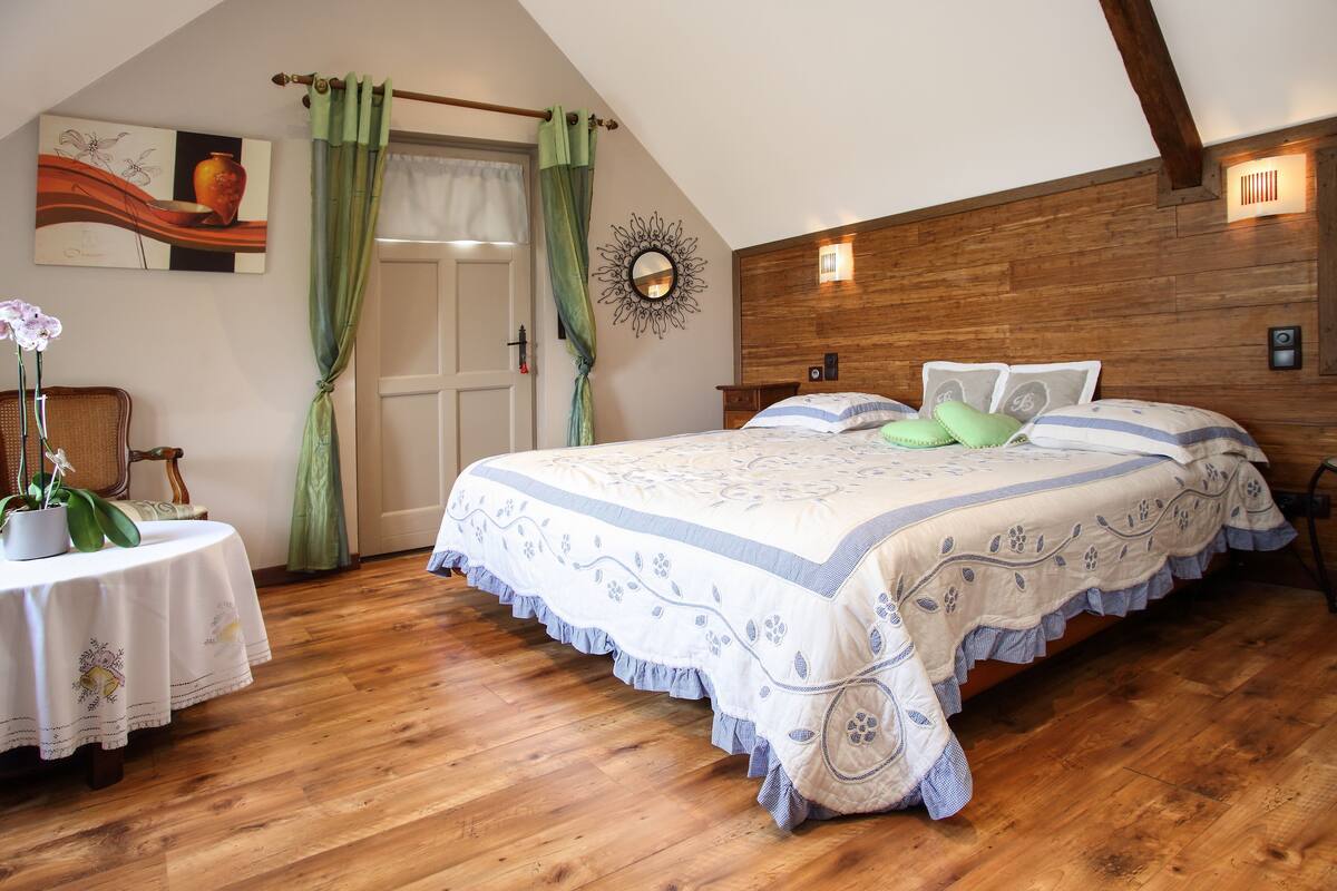Une chambre avec un plafond en bois, un lit recouvert d’une couvre-lit à motifs floraux, une table de chevet ornée d’une plante en pot et un miroir fixé au mur.