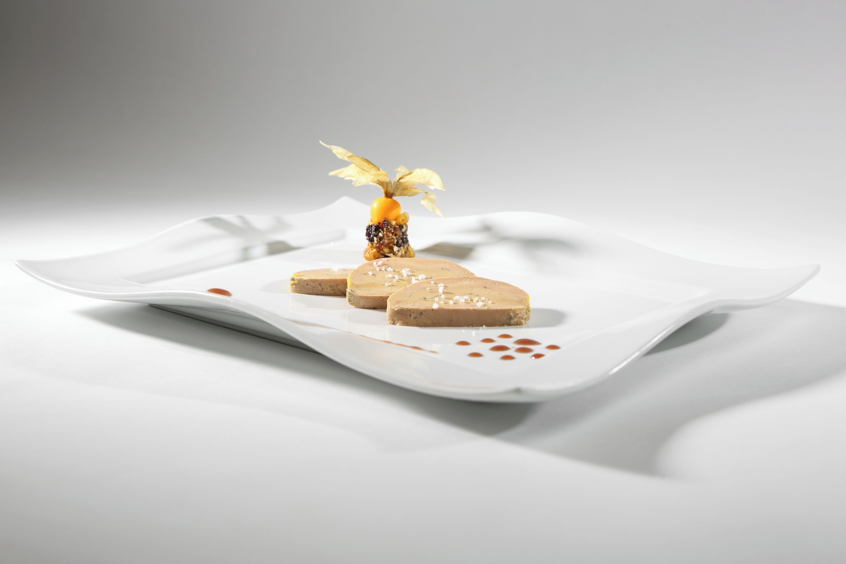 Un piatto bianco con un dessert, un elemento centrale sormontato da un frutto giallo e una foglia, circondato da piccoli pezzi.