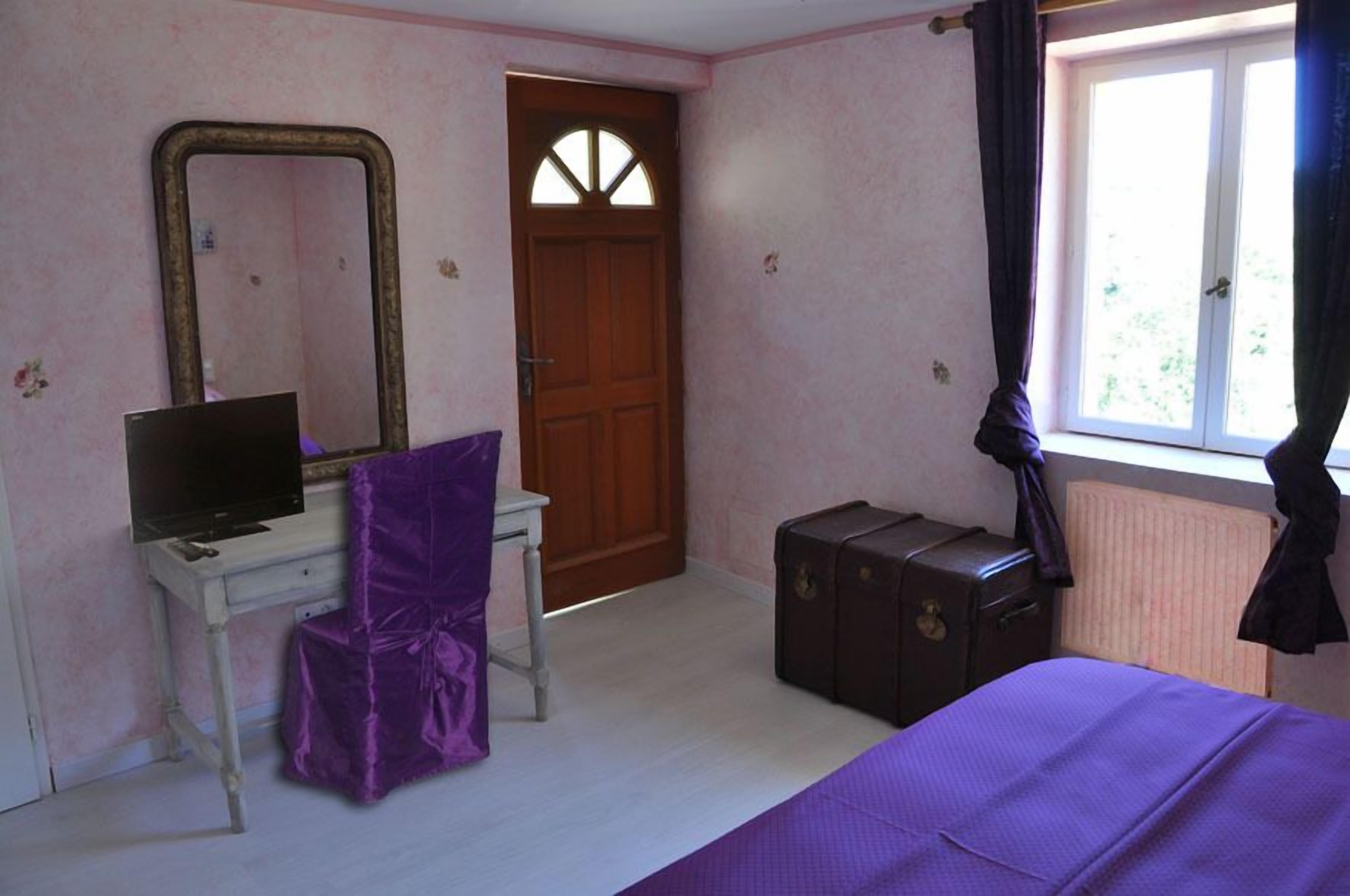 Un dormitorio con una cama, un escritorio con computadora, un tocador, un espejo, una puerta y una ventana con cortinas.