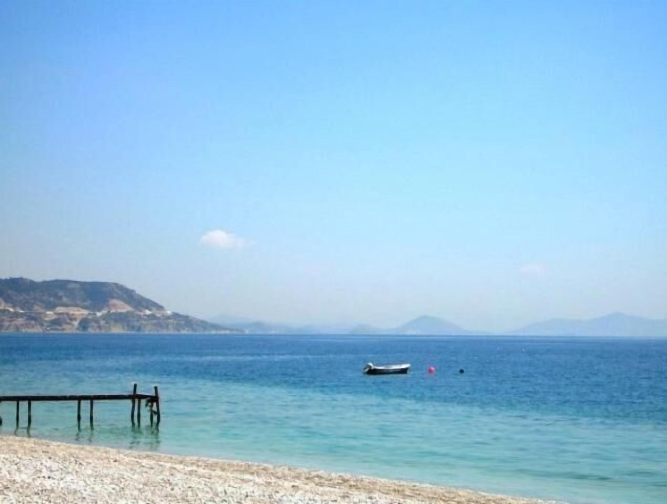 A beach with a wooden pier extending into calm waters, a small boat, and distant hills under a clear sky.