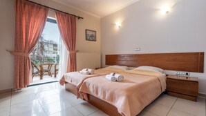 A double bed with a wooden headboard, a bedside table with a lamp, and a view of a balcony with a chair. - J Studios (Megara)