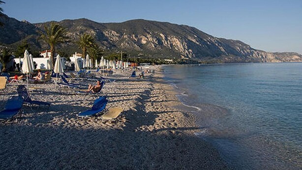 A beach with umbrellas and lounge chairs, a calm sea, and a mountainous backdrop. - J Studios (Megara)