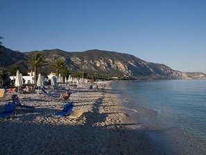 Una playa con sombrillas y sillas de descanso, un mar tranquilo y montañas al fondo.