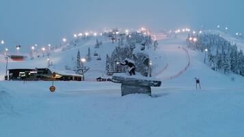 A snow-covered ski slope with a snowboarder performing a trick on a rail, surrounded by ski lifts and illuminated by artificial lights.