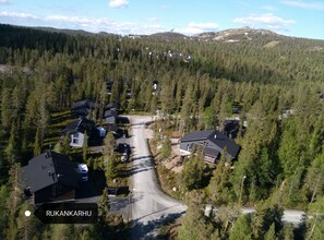 An aerial view of a residential area with houses surrounded by dense forest, a winding road, and a hilly landscape in the background.