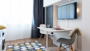A compact room with a white desk, a chair, a TV mounted on the wall, and a patterned rug. - WE Apartments (Warsaw)