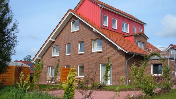 Ein zweigeschossiges Backsteinhaus mit rotem Dach, zahlreichen Fenstern und einem kleinen Garten mit Rasen und Pflanzen.