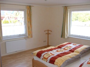 Un dormitorio con cama, radiador, perchero de madera y dos ventanas con cortinas.