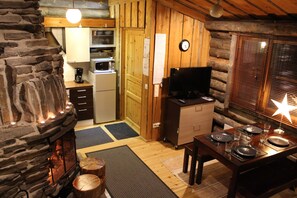 A cozy cabin interior with a stone fireplace, wooden walls, a kitchen area, and a dining table set for four.