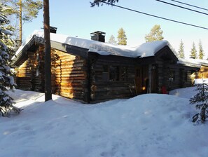 Una cabaña de troncos con techo nevado, rodeada de nieve y árboles.