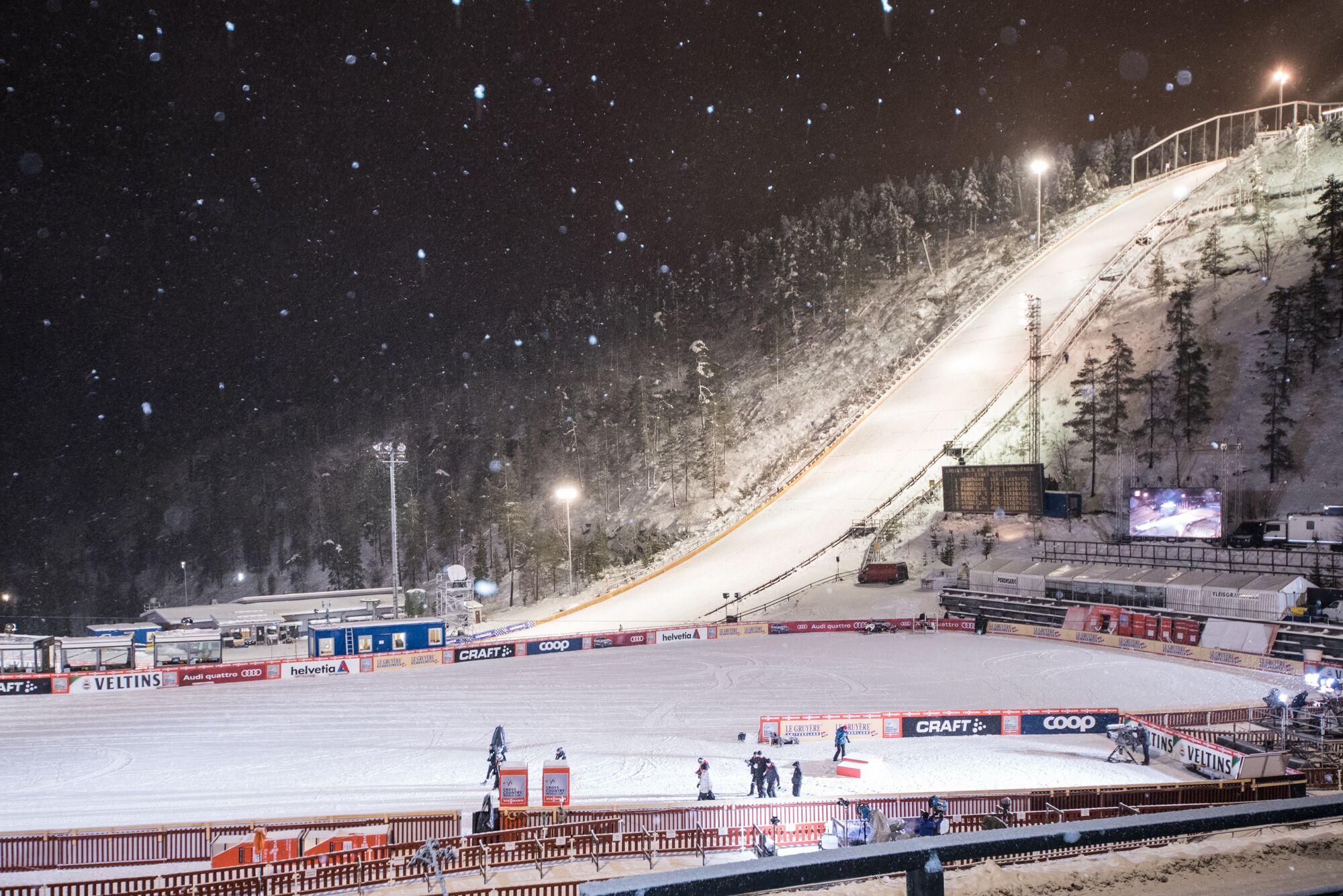 Pista de esqui coberta com neve, arquibancadas para espectadores e uma tela grande exibindo eventos.