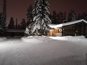 Una cabaña de troncos con techo cubierto de nieve y una entrada iluminada, rodeada de nieve y árboles.