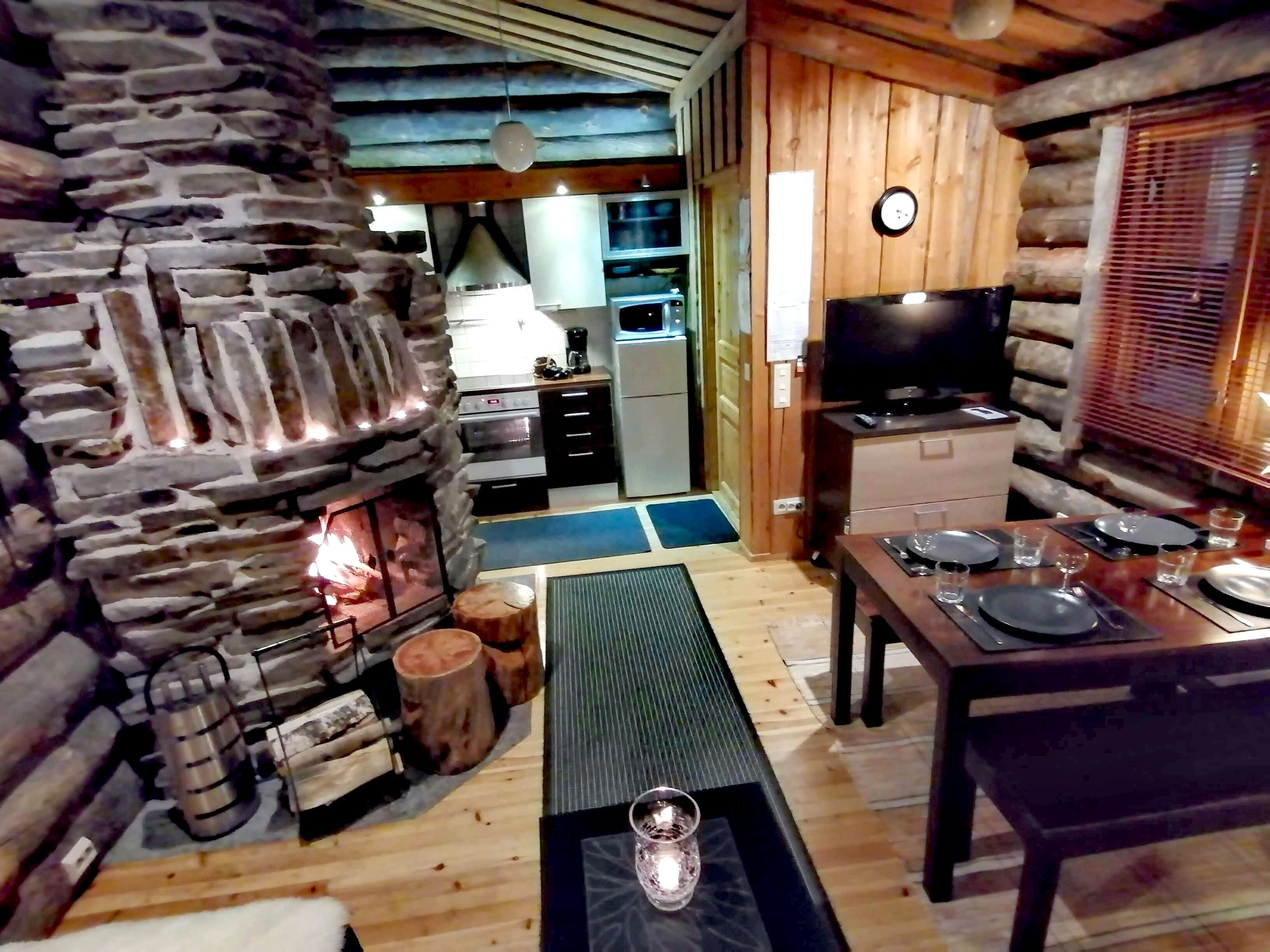 Un acogedor interior de cabaña con chimenea de piedra, una mesa de comedor de madera dispuesta para dos personas y una zona de cocina con electrodomésticos modernos.