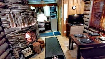 Un acogedor interior de cabaña con chimenea de piedra, una mesa de comedor de madera dispuesta para dos personas y una zona de cocina con electrodomésticos modernos.