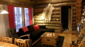 Interior de una cabaña de troncos con un sofá, sillas de madera, una mesita y una chimenea.