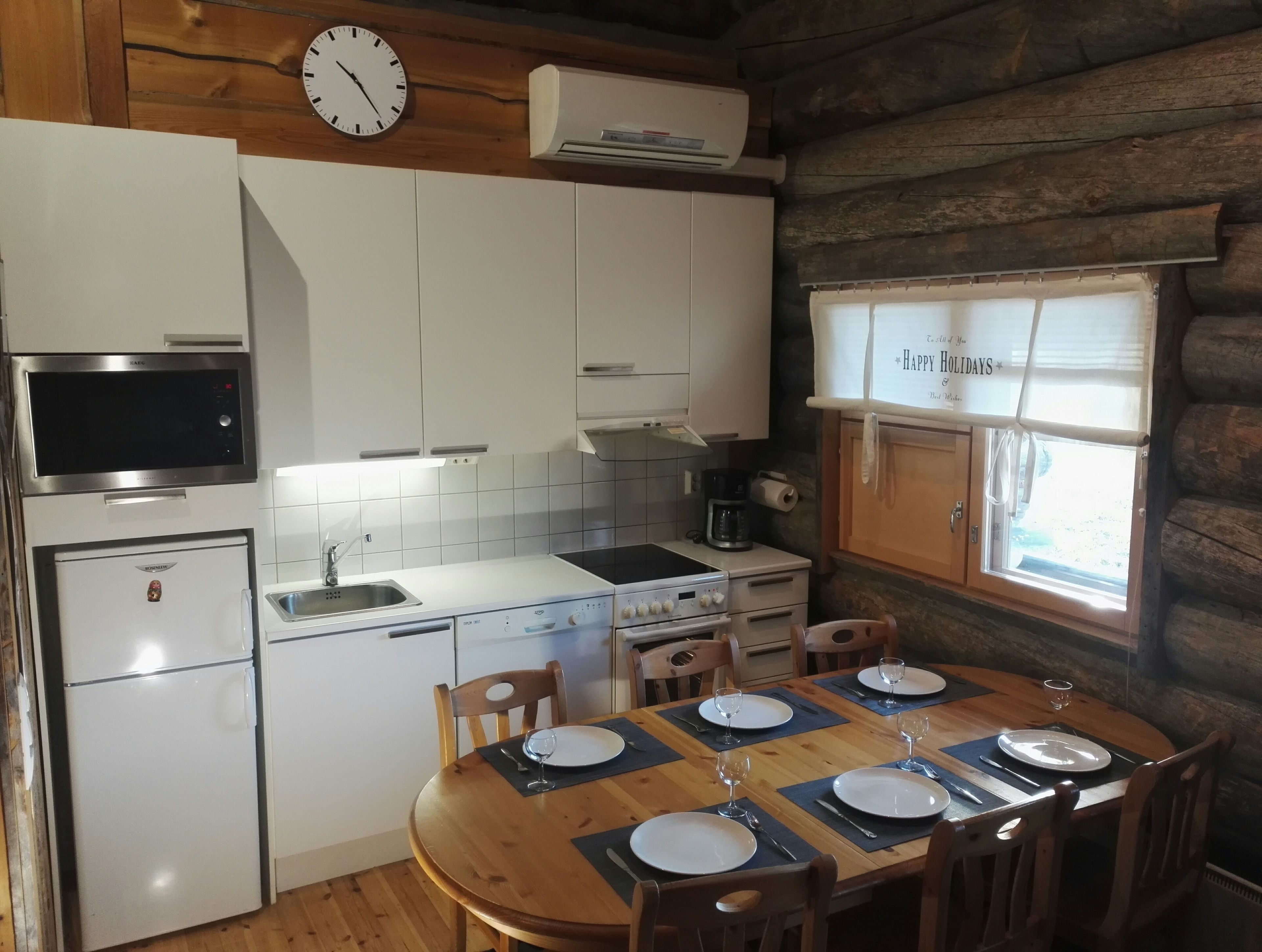 Een kleine keuken met een magnetron, fornuis en spoelbak. Een eettafel is gedekt voor vier personen, met borden, glazen en bestek. Op de muur hangt een klok, en er is een raam met gordijn.
