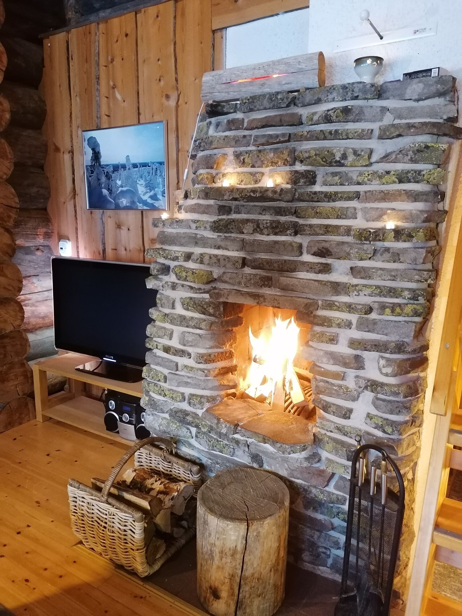 Une cheminée en pierre avec un feu de bois, un téléviseur posé sur un meuble en bois et un panier de bûches.