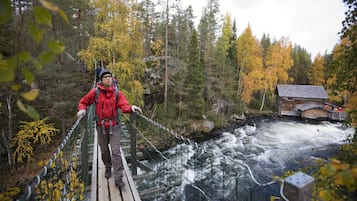 A person crossing a suspension bridge over a river in a forested area with a cabin in the background.