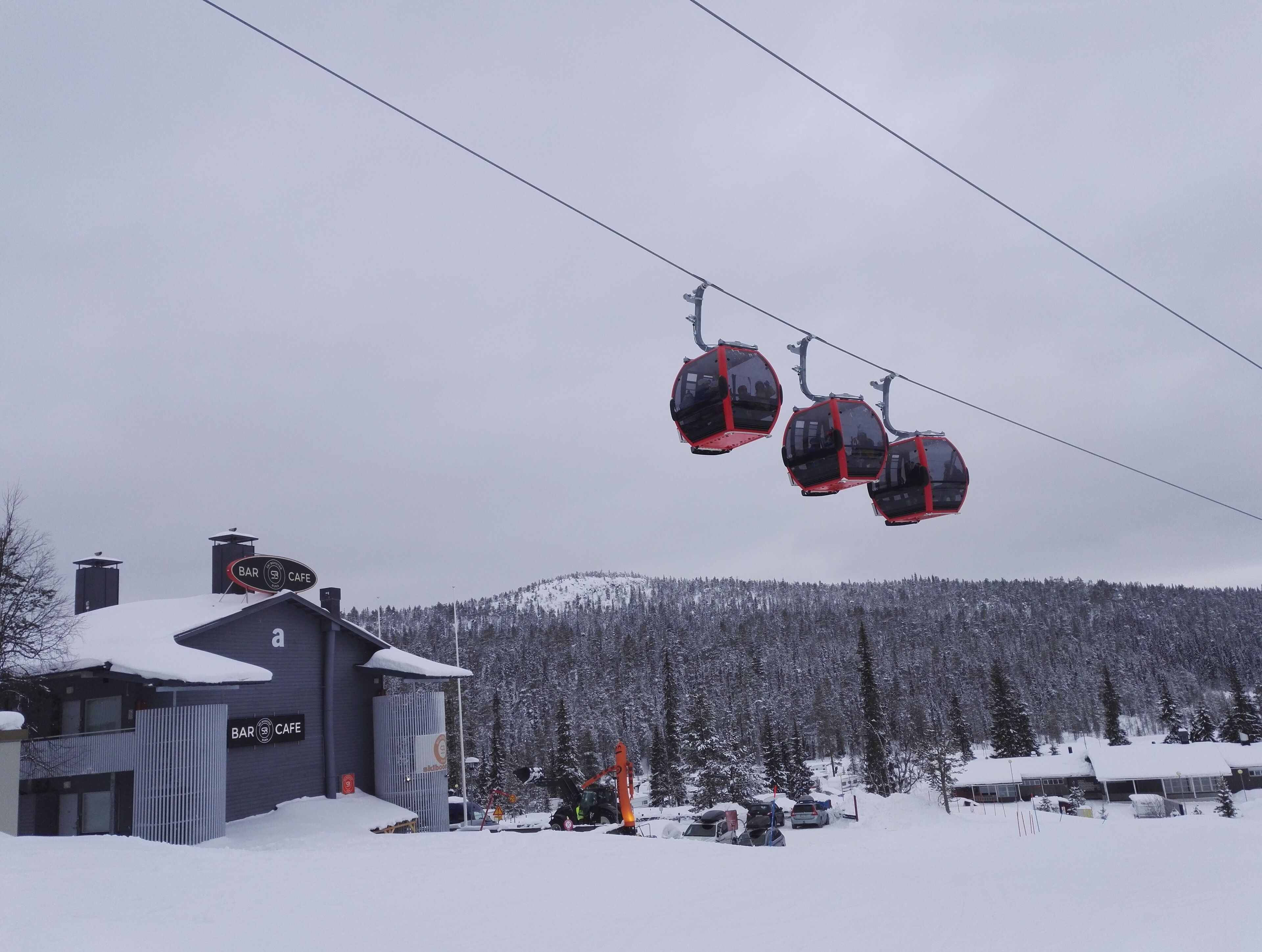A ski lift with cabins, a building with a sign for a bar and cafe, and a snowy mountain in the background.