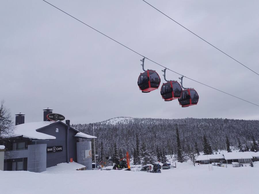 A ski lift with cabins, a building with a sign for a bar and cafe, and a snowy mountain in the background.