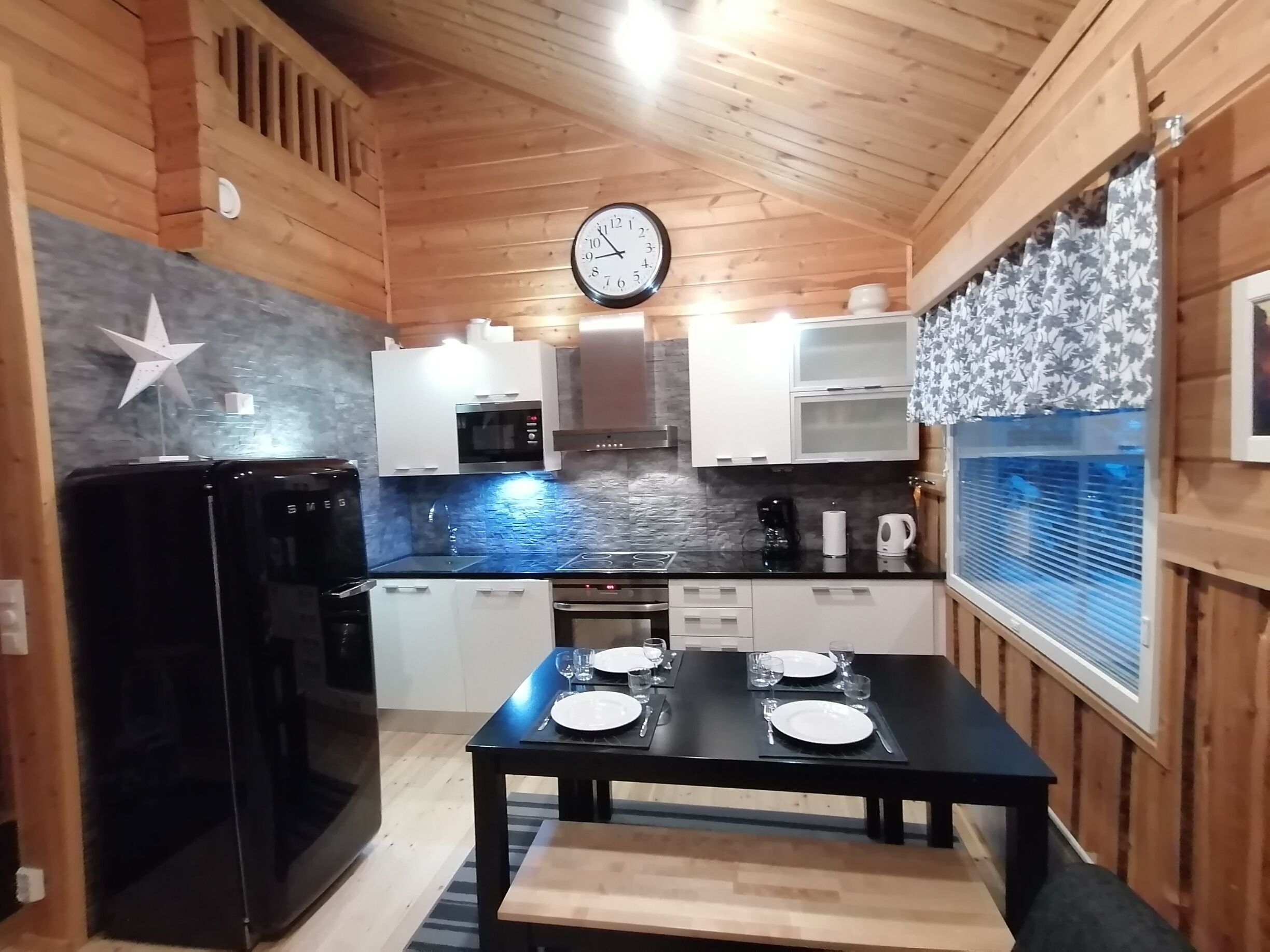 A compact kitchen with wooden walls, a black refrigerator, a microwave, and a clock on the wall.
