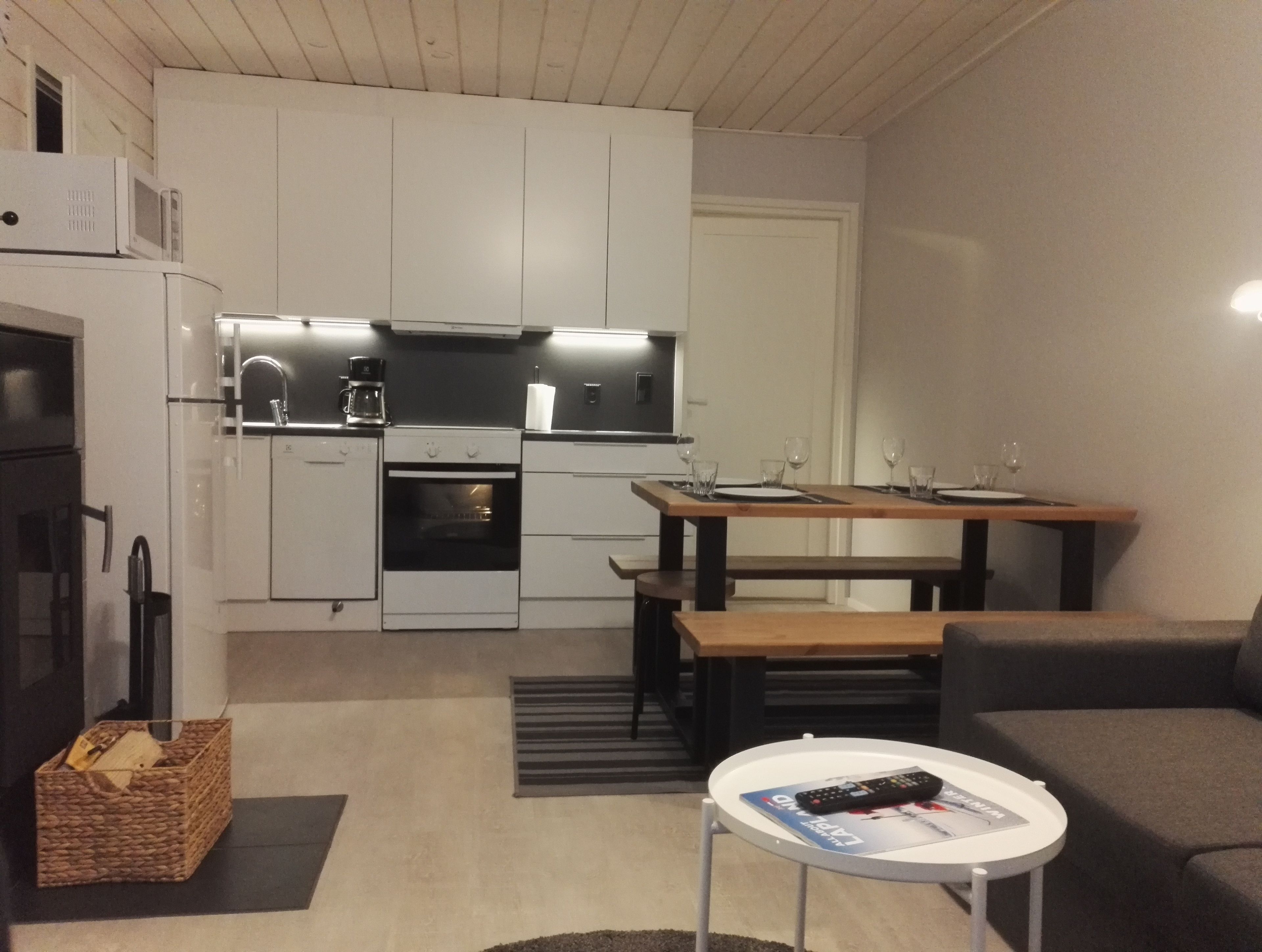 Kompakti keittiö, jossa on valkoiset kaapit, sisäänrakennettu uuni ja mikroaaltouuni. Siellä on myös ruokailutila, jossa on pöytä ja tuolit, sekä pieni pyöreä pöytä, jonka päällä on lehti ja kaukosäädin.