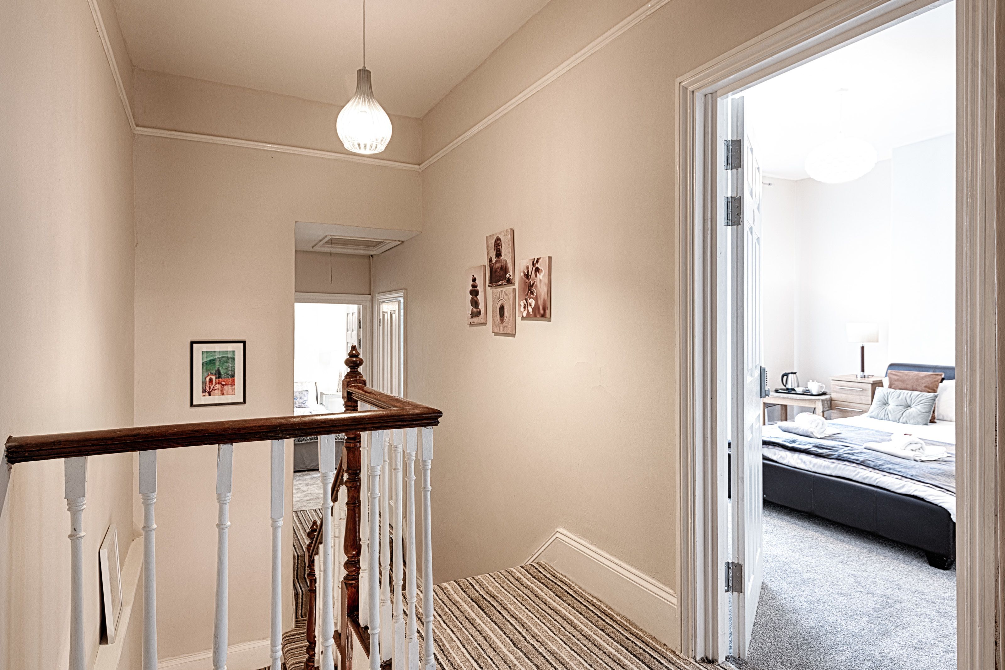 A hallway leading to a bedroom with a bed, a nightstand, and a lamp. There is a staircase with a wooden handrail and white balusters.