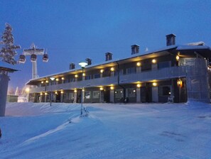A multi-story hotel with a snow-covered ground, a ski lift in the background, and a balcony with lights. - Vuosselin Helmi (Kuusamo)
