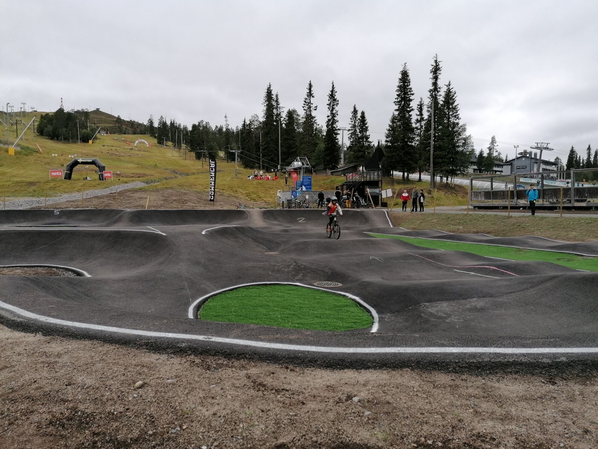 A BMX track with a cyclist in action, surrounded by spectators and a hilly landscape with trees and a ski lift in the background.